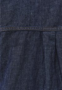 Mørkeblåt denimstof med synlige vævninger og en vandret søm syet med brunt tråd nær toppen.