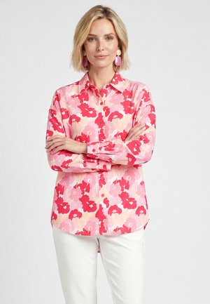 Vrouw met kort blond haar, die een roze en rode bloementrui met knopen draagt, witte broek en opvallende roze oorbellen, armen gekruist.