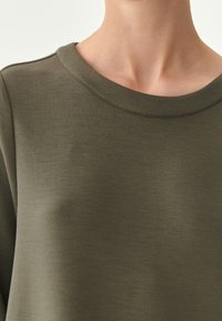 Felpa verde oliva con scollatura rotonda. Tessuto morbido e liscio con una leggera lucentezza; presenta dettagli di cucitura minimi lungo il colletto.