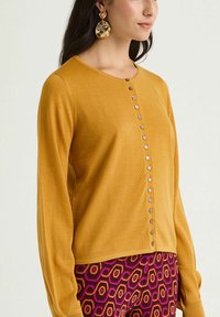 Cardigan jaune moutarde à manches longues, col rond et boutonnage à l'avant. Présente une texture douce et une finition lisse.