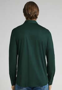 Hombre visto desde atrás, con una camisa de manga larga verde oscuro y jeans azules, contra un fondo gris claro liso.