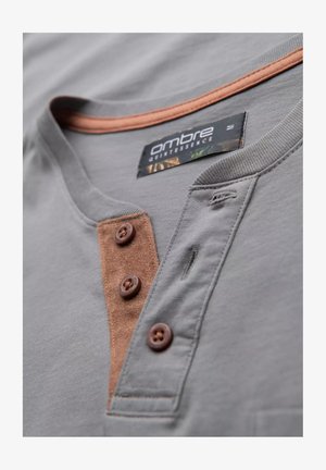 Grijs shirt met korte mouwen met bruin suède accent en drie bruine knopen. Heeft een ronde hals met contrasterende oranje rand.