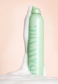 Ljust grön cylindrisk flaska märkt "IDA WARG Hydrating Shower Mousse" delvis nedsänkt i vit skum mot en mjuk rosa bakgrund.