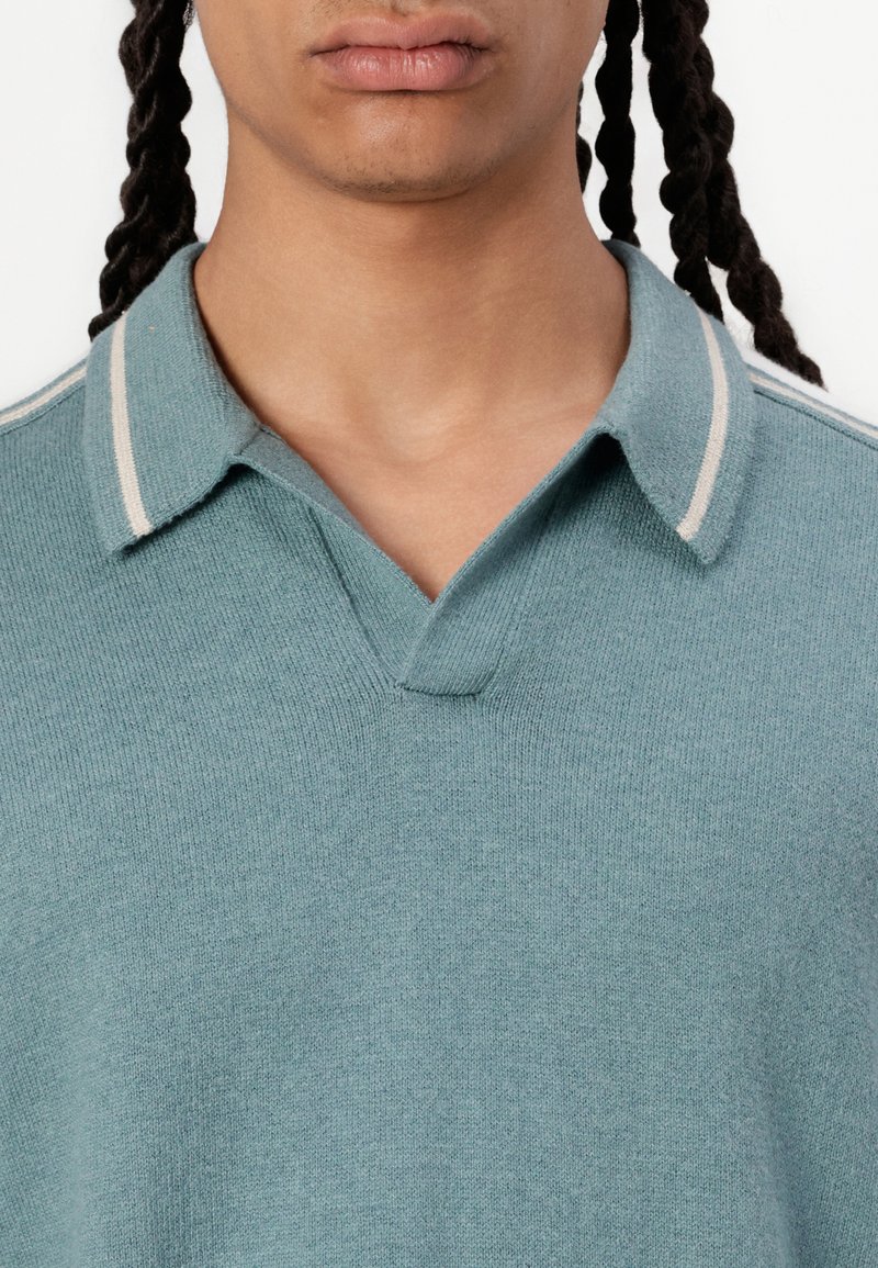 Polo en teal clair avec un col en V, fabriqué à partir d'un tissu texturé. Doté d'un col classique avec des rayures blanches contrastantes.