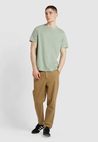 T-shirt verde chiaro a maniche corte con collo a giro, dettagli ricamati piccoli, abbinato a pantaloni larghi color beige e sneakers nere.