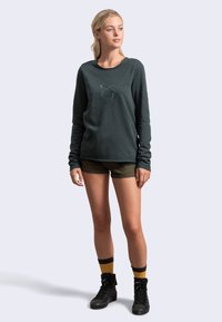 Felpa a maniche lunghe verde scuro con un design astratto blu chiaro sul petto, abbinata a pantaloni corti olive e scarponcini neri con lacci.