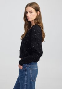 Donna con lunghi capelli castani che indossa un maglione nero a macchie e jeans blu, in piedi di lato con una mano nella tasca contro uno sfondo semplice.