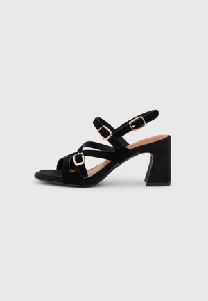 Black suede high-heeled sandals with crisscross straps, gold square buckles, open toe, and a chunky heel. Smooth texture and minimal design.