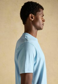Jeune homme aux cheveux courts et bouclés, portant une chemise à manches courtes bleu clair, debout de profil devant un fond beige uni.