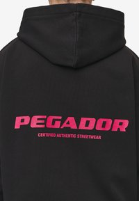 Black hoodie with a large pink graphic text reading "PEGADOR" and "CERTIFIED AUTHENTIC STREETWEAR" on the back. Soft texture, hooded design.