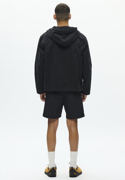 Veste noire à capuche avec une coupe décontractée, associée à un short noir. Le modèle porte des chaussettes blanches et des chaussures de sport jaunes et noires.