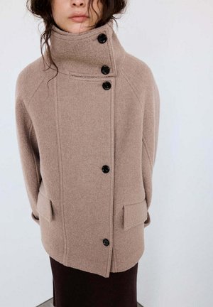 Femme portant un manteau en laine beige avec un col montant et quatre boutons noirs, debout devant un fond clair uni.