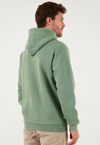 Vihreä huppari pehmeästä kankaasta, jossa on kengurutasku, resorit hihansuissa ja helmassa, suuri huppu ja sileä pinta.