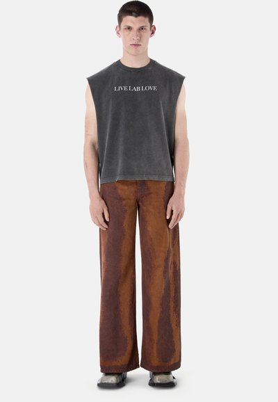 Junger Mann steht, trägt ärmelloses schwarzes Oberteil mit dem Schriftzug "LIVE LAB LOVE" und weit geschnittene braune Batik-Hosen mit schwarzen Schuhen.