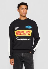 Svart sweatshirt med flerfärgade graphics, med gul och röd text "REPLAY" och vita sidopaneler. Tillverkad av mjukt tyg med rundad halsringning.
