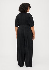 Curly-haired woman in a black pleated jumpsuit, viewed from the back, stands against a plain white background.