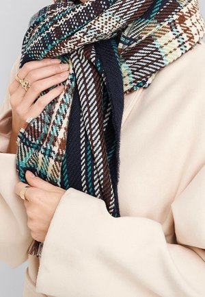 Persoon draagt een beige jas en houdt een dikke, meerkleurige geruite sjaal met bruine, petrolblauwe, zwarte en witte patronen om de nek.