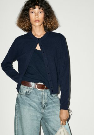 Donna con capelli ricci indossa un cardigan blu navy sopra un top nero, jeans azzurri con cintura marrone e tiene una borsa chiara.