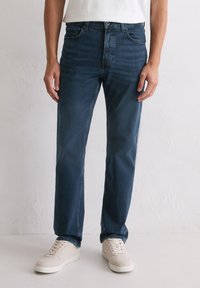 Homme portant un jean droit bleu foncé et des baskets beige clair, adossé à un mur blanc uni.