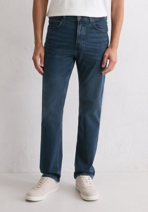 Mężczyzna w ciemnoniebieskich jeansach o prostym kroju i jasnobeżowych trampkach stoi przy gładkiej białej ścianie.