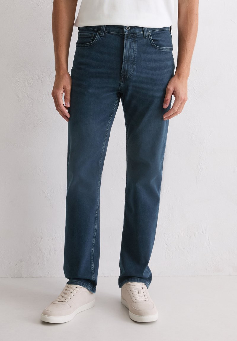 Homme portant un jean droit bleu foncé et des baskets beige clair, adossé à un mur blanc uni.