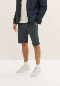 Kurze, weiße Hemd unter einer marineblauen gesteppte Jacke, kombiniert mit gemusterten marineblauen Shorts und weißen Sneakers mit hellen Sohlen.