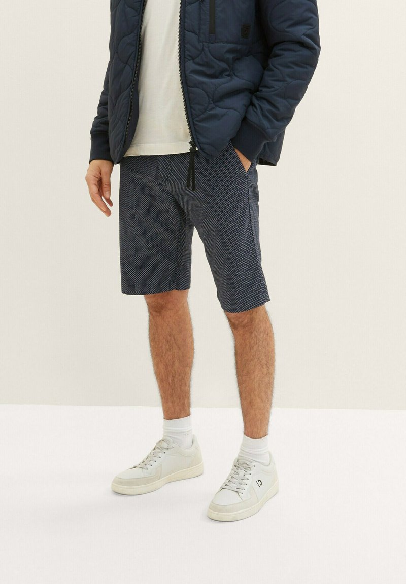 Kurze, weiße Hemd unter einer marineblauen gesteppte Jacke, kombiniert mit gemusterten marineblauen Shorts und weißen Sneakers mit hellen Sohlen.