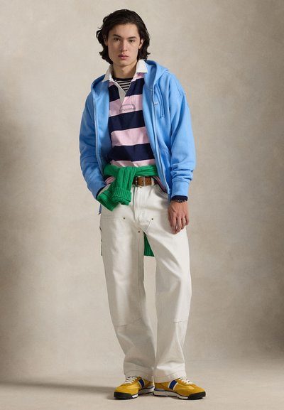 Zils džemperis ar kapuci virs rozā un tumši zilām svītrām ar polo krekliņu, baltas bikses un dzelteni sporta apavi ar ziliem akcentiem. Zaļš džemperis sasiets ap vidukli.