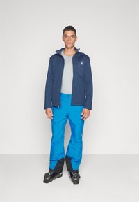 Blaue Skihosen, graues Shirt und eine marineblaue Jacke mit einem Spinnen-Logo. Die Hosen haben schwarze Akzente an den Säumen. Hergestellt aus wasserfesten Materialien.