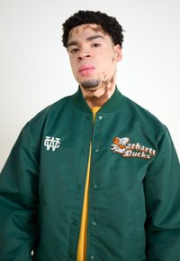 Grøn bomberjakke med knapper, med et broderet "Carhartt Ducks" logo og en hvid grafik. Båret over en gul skjorte.