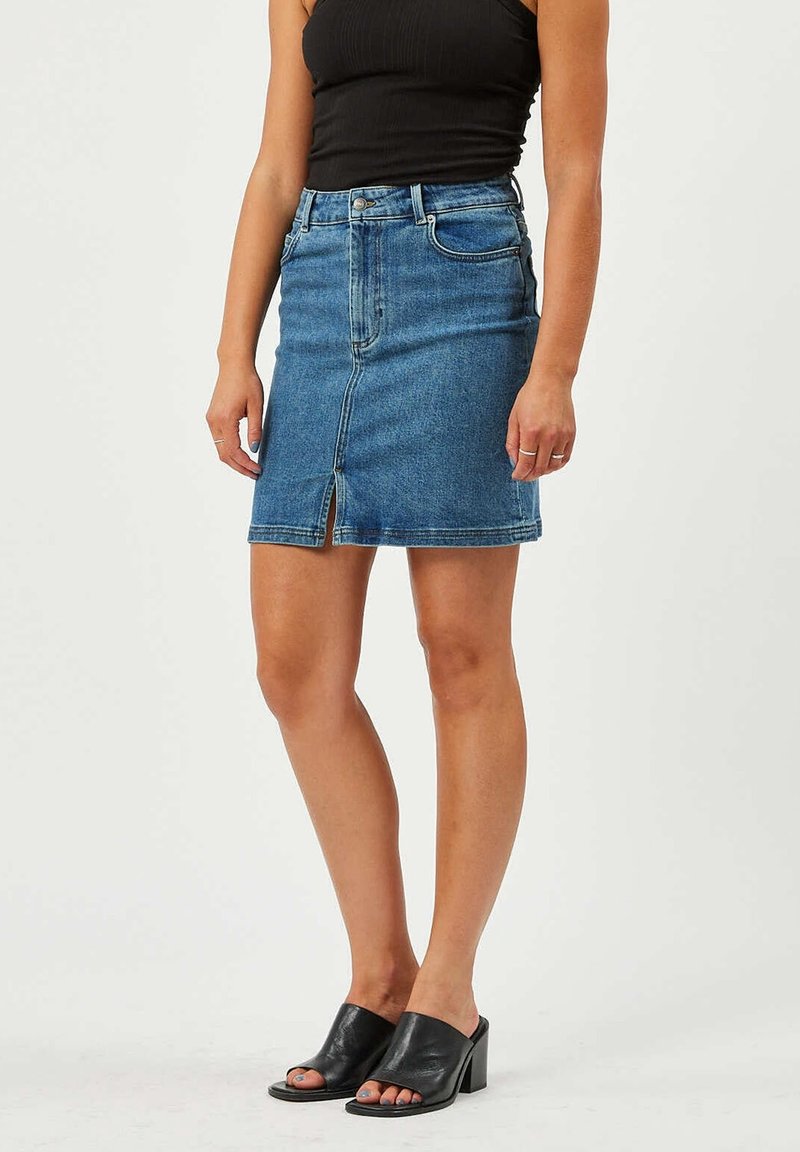 Femme portant une jupe en denim bleu avec une fente frontale, un haut sans manches noir, et des mules ouvertes à talons noirs, se tenant sur un fond uni.