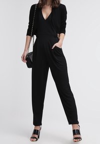 Schwarzer Jumpsuit mit tiefem V-Ausschnitt, tailliertem Bund und weitem Bein, kombiniert mit einer enganliegenden schwarzen Strickjacke. Glattes, leichtes Material.