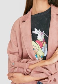 Veste en suédine rose à coupe décontractée, avec un col noté et des poignets boutonnés. En dessous, un T-shirt noir à motif graphique avec des personnages de dessin animé.