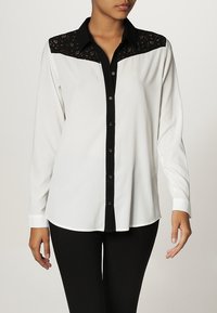 Blouse blanche avec empiècement en dentelle noire et fermeture boutonnée devant. Manches longues, col noir et poignets. Tissu lisse avec un léger éclat.