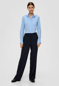Hellblaues Button-down-Hemd mit langen Ärmeln, kombiniert mit marineblauen Weitbeinhosen und schwarzen Schuhen, vor einem schlichten grauen Hintergrund.