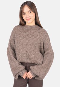 Brauner, grob gestrickter Pullover mit hohem Kragen und übergroßen Ärmeln. Weiche Textur mit gerippten Bündchen, getragen über einer dunklen Hose.