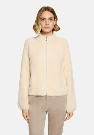 Femme portant une veste crème en tissu duveteux zippée à manches longues et un pantalon beige, debout devant un fond clair uni.