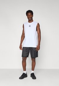 Chemise sans manches blanche avec un logo noir NY, associée à un short en mesh noir et des baskets noires. Arrière-plan gris neutre.
