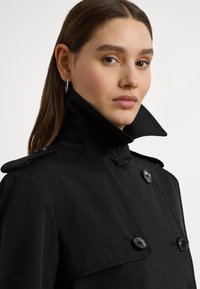 Black trench coat featuring a high collar, double-breasted design, button accents, and a smooth fabric finish.
