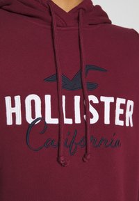 Felpa con cappuccio color marrone scuro con scritta bianca "HOLLISTER" e scritta in corsivo blu "California". Presenta una tasca a marsupio e cappuccio con coulisse.