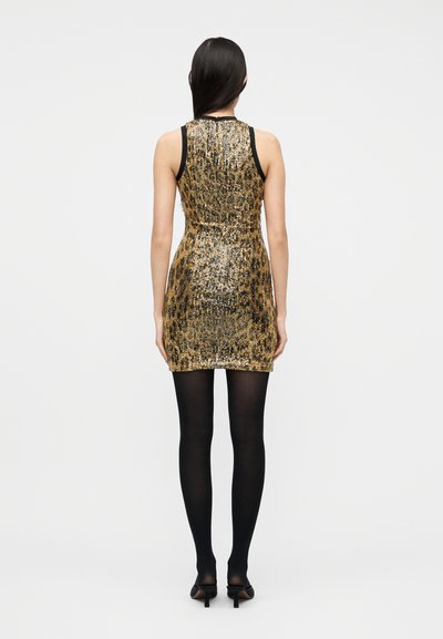 Γυναίκα με μίνι φόρεμα χωρίς μανίκια, χρυσό και μαύρο με παγιέτες, μαύρο καλσόν και μαύρα παπούτσια με τακούνι, στέκεται γυρισμένη από την πλάτη σε απλό φόντο.