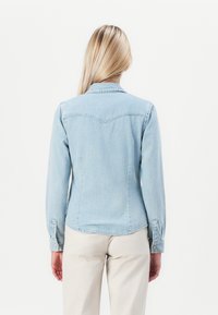 Chaqueta de denim azul claro con mangas largas, cuello clásico y puños abotonados. Dobladillo curvado y sutil detalle de yugo en la parte posterior. Textura lisa.