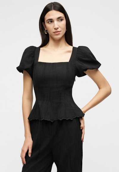 Γυναίκα που φοράει εφαρμοστό μαύρο τοπ με φουσκωτά μανίκια, τετράγωνη λαιμόκοψη, με τελείωμα πεπλούμ, σε συνδυασμό με μαύρο παντελόνι, στέκεται με το ένα χέρι στη μέση.