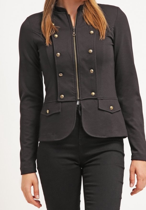 Femme portant une veste noire ajustée avec des boutons dorés, fermeture éclair devant, poches à rabat et pantalon noir assorti.