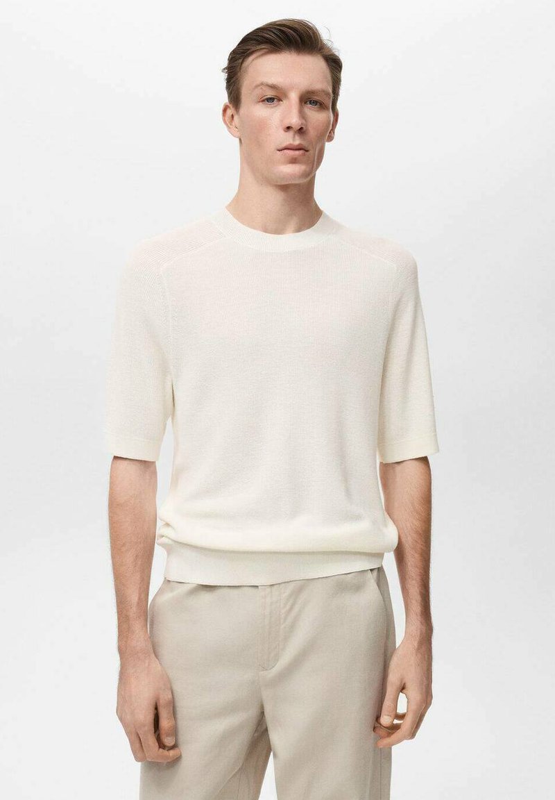 Pleteni pulover s kratkimi rokavi v off-white barvi, ki ima okroglo ovratnik, rebrasto teksturo in nekoliko prilegajoč dizajn, v kombinaciji s svetlobezž pantami.