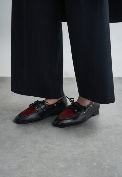 Black leather loafers with burgundy felt accent, lace-up design, gold eyelets, low profile, worn with dark pinstripe trousers.