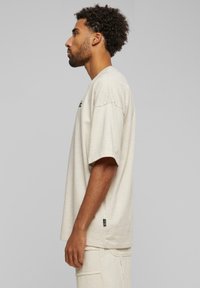 Beige oversized tričko vyrobené z jemného materiálu, s krátkymi rukávmi, okrúhlym výstrihom a malým logom na hrudi.