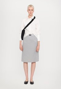 Fustă gri până la genunchi, cu o textură netedă și un petic cu logo negru, asortată cu o cămașă albă cu nasturi și pantofi negri cu talpă joasă.