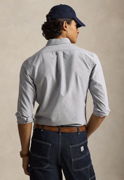 Man wearing light gray button-down shirt with sleeves rolled up, dark denim pants, brown belt, navy cap, shown from the back against plain background.