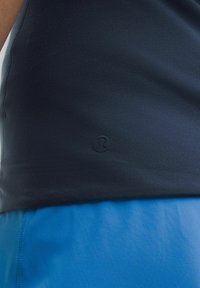 Tamsiai mėlynas sportinis maikutis su glotniu paviršiumi, su mažyte juoda logotipu apačioje, derinamas su šviesiai mėlynomis šortais.